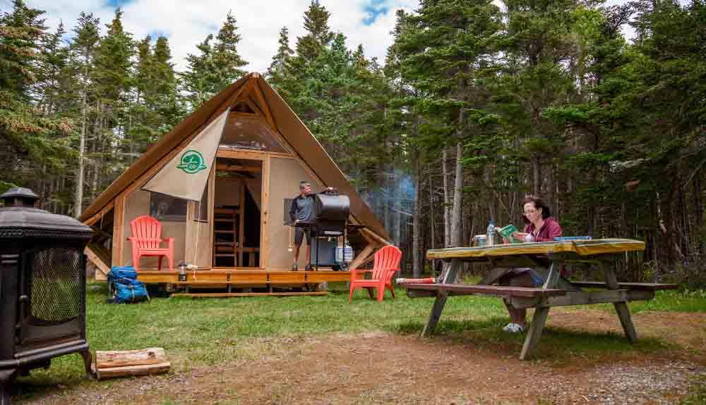 A couple sits at a picnic table in front of a tent in the woods.