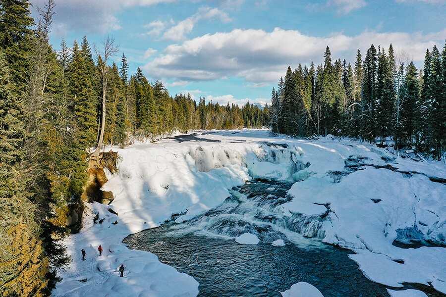A partially frozen waterfall with icy banks surrounded by a coniferous forest, featuring a group of people observing near the edge.