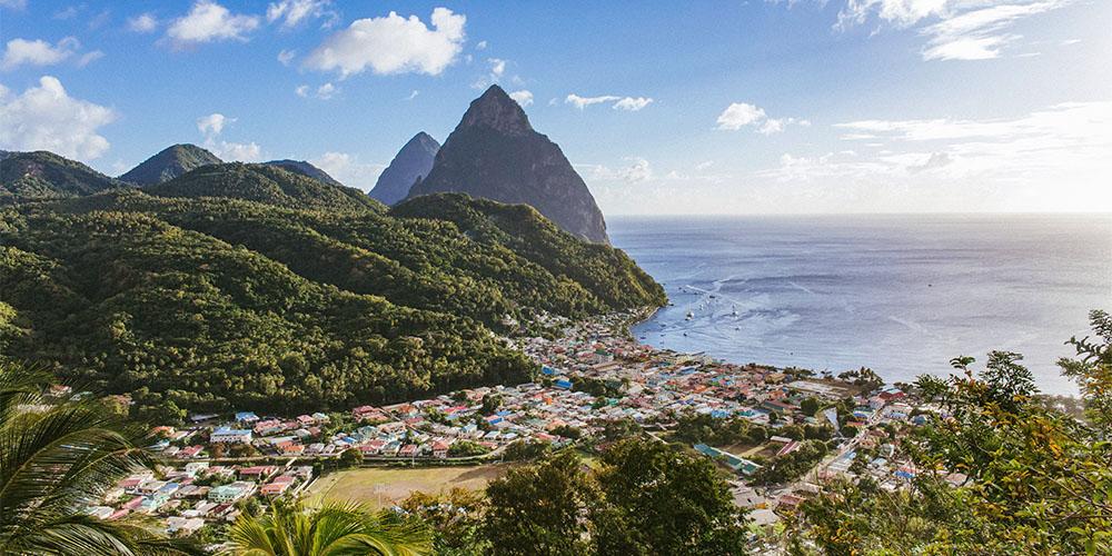 A coastal town nestled between lush green hills and the ocean, with towering peaks in the background.