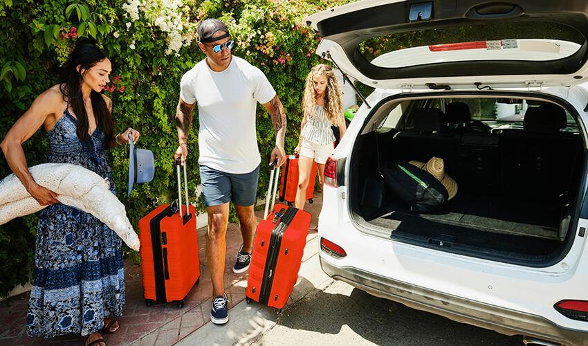 Family unloading luggage from a car trunk into a sunny driveway.