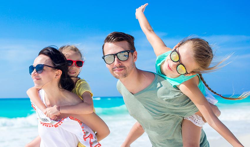 A family enjoying a sunny day at the beach, with two children on their parents' backs.