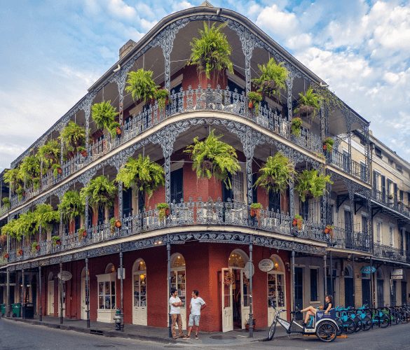 An ornate two-story building with wrought-iron balconies, adorned with green plants, on a street corner in a historic district, with pedestrians and a parked pedicab nearby.