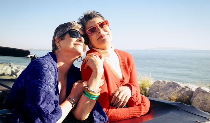 Two women sitting on the back of a car with the ocean in the background.