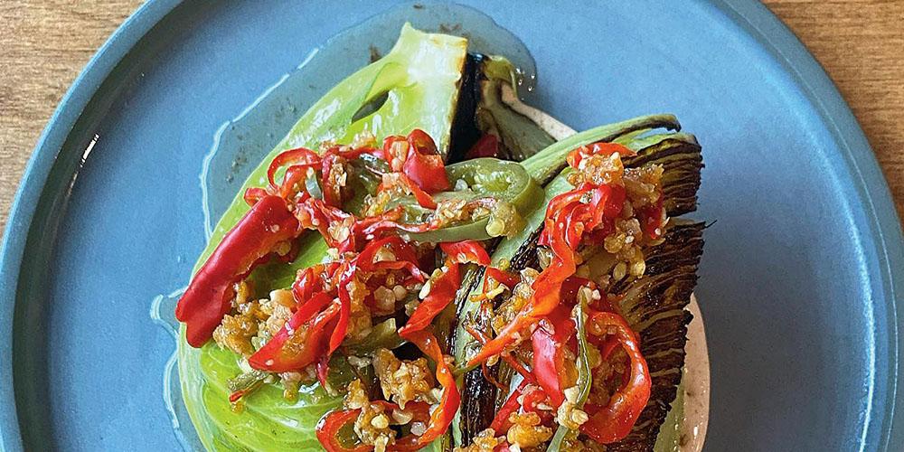 Grilled bok choy topped with chili peppers and a savory garnish, served on a blue plate.