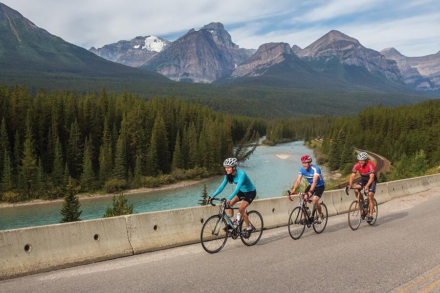 Three people riding bicycles on a road with mountains in the background.