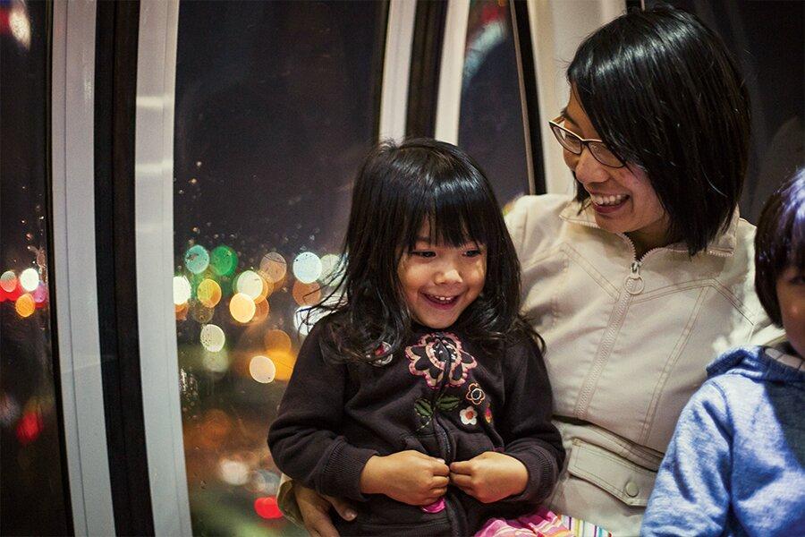 A woman smiling at a joyful young girl in a dimly lit space, possibly a vehicle, with blurred city lights in the background.