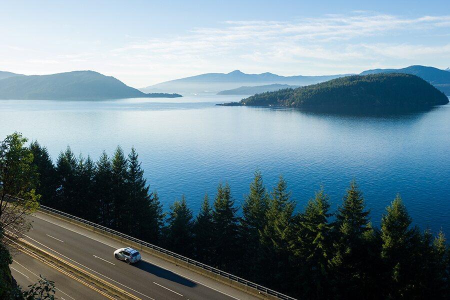A car driving along a coastal road with a view of a serene blue lake and forested hills under a clear sky.