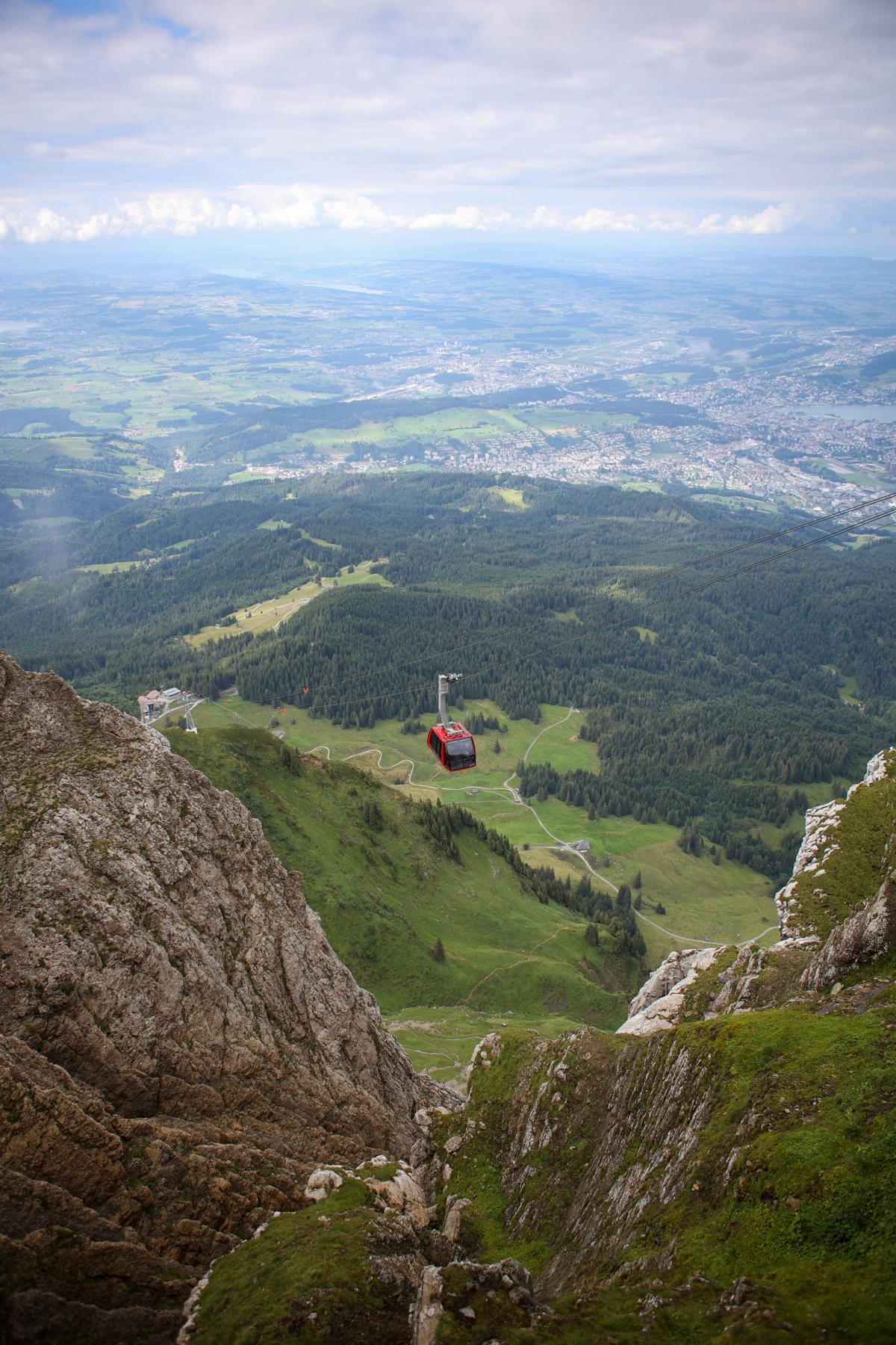 An overhead shot of a red cable car making its way up from one mountain to another. Below and behind it are green fields with dense trees.