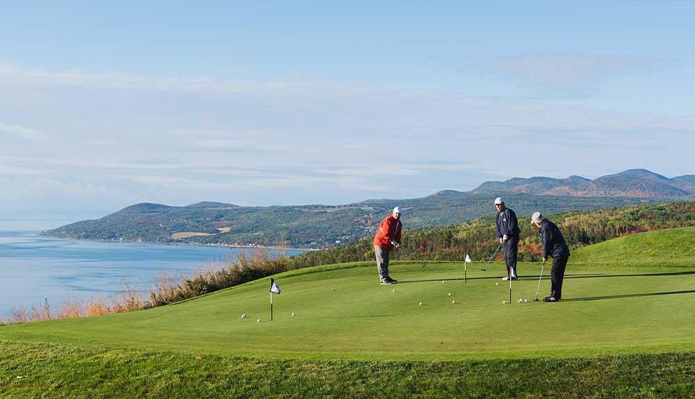 Three men playing golf on a hill overlooking the ocean.