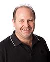A smiling man with stubble wearing a black polo shirt against a white background.