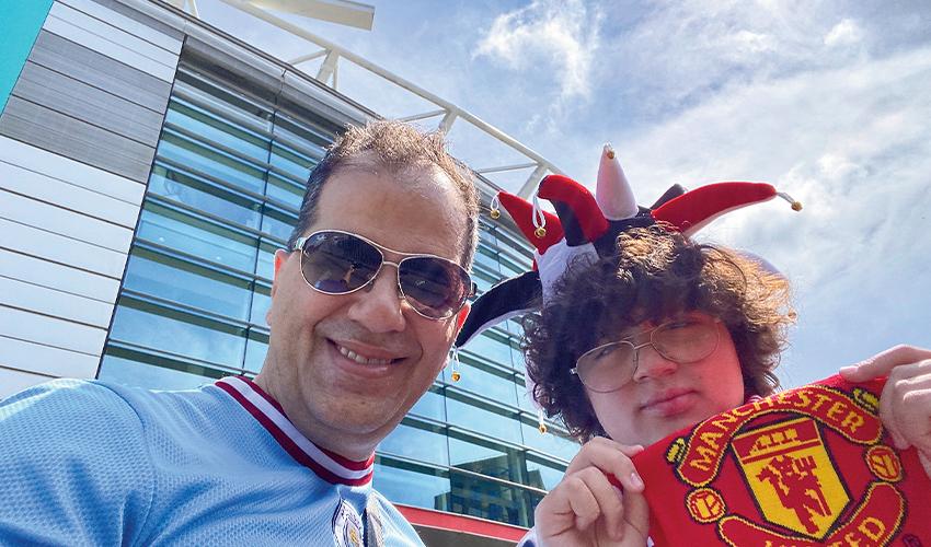 Two soccer fans posing in the sunshine, with one wearing a jester hat and holding a manchester united scarf.
