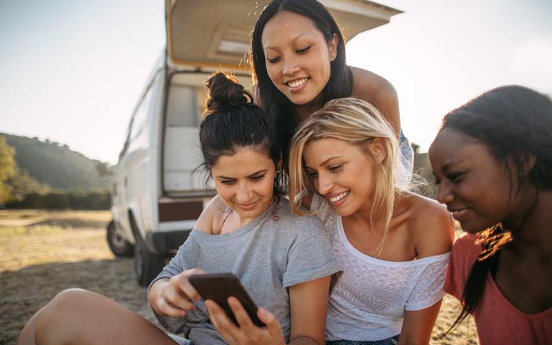 Four woman sitting outside looking at a phone behind a parked car in a wilderness