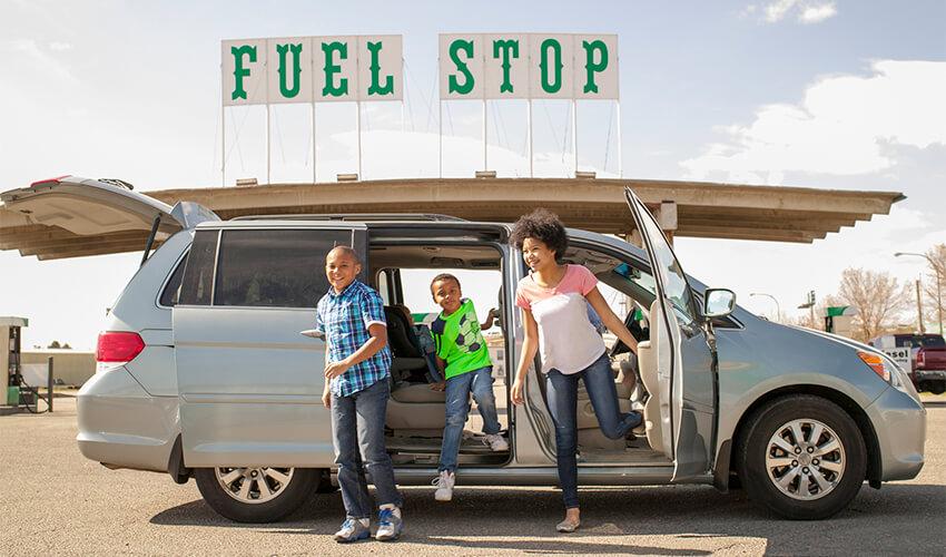 A family getting out of their minivan at a fuel stop on a sunny day.