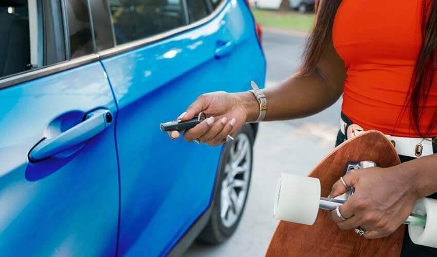 A woman holding a skateboard next to a blue car.