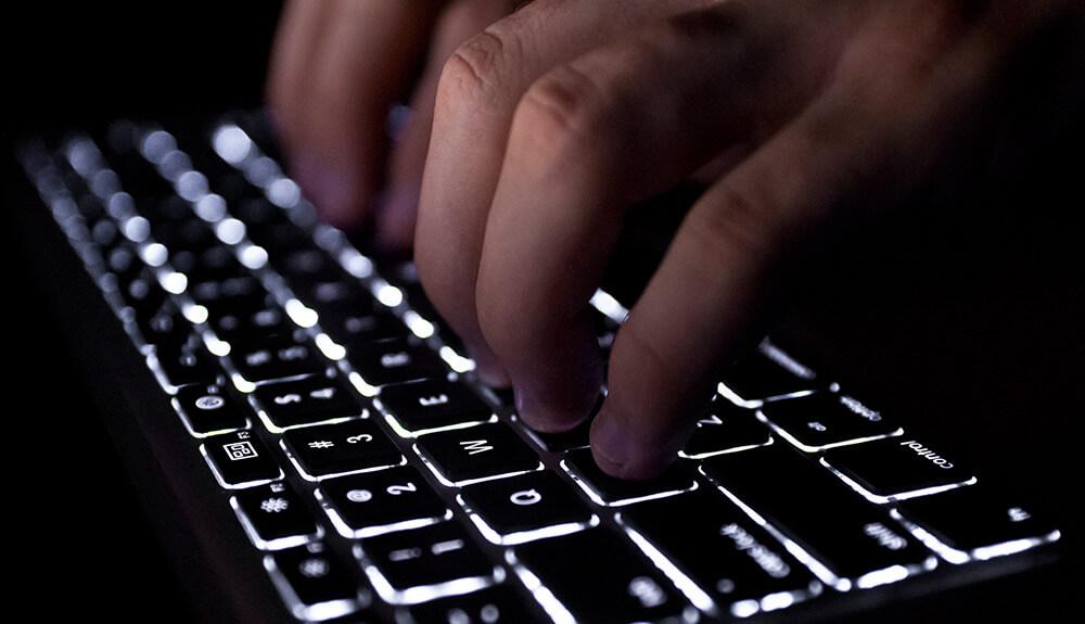 Fingers typing on a backlit laptop keyboard in low-light conditions.
