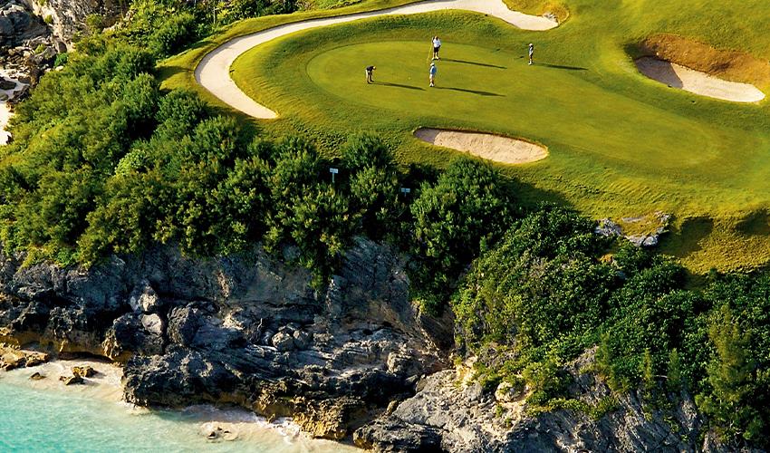 Aerial view of golfers on a scenic coastal golf course with undulating green and adjacent rocky shoreline.