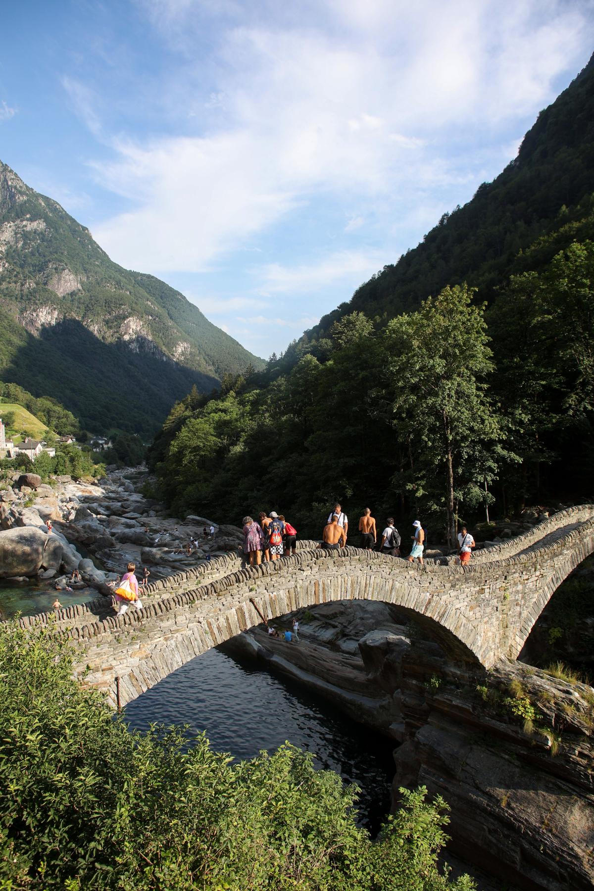 A stone bridge between mountains covered in bushes and trees with people standing all along it.