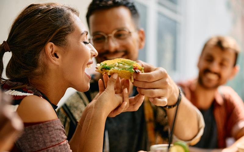 Man feeding his woman a taco with friends around