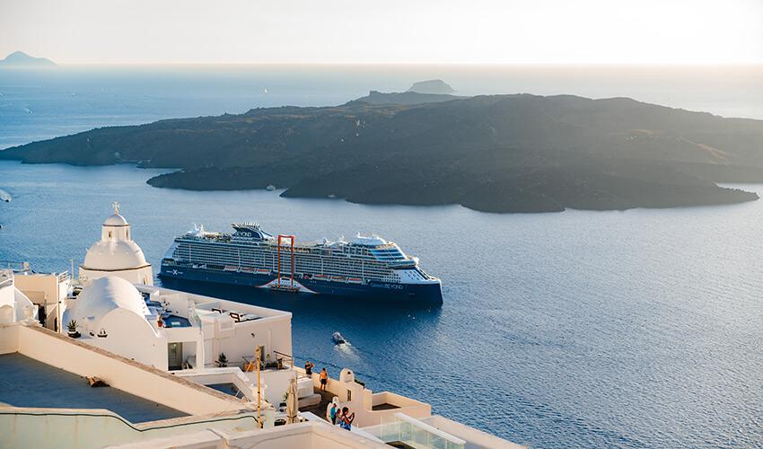 A cruise ship anchored near a coastline with white buildings and a view of the sea during sunset.