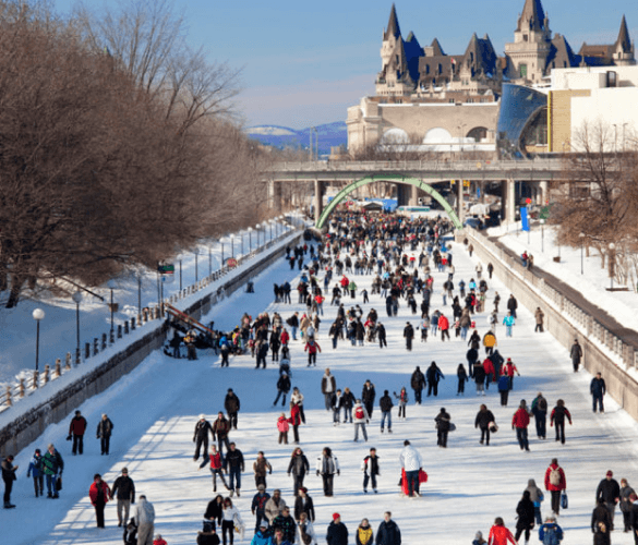 Crowds of people ice skating on a wide outdoor rink in a city setting with a historical building in the background.